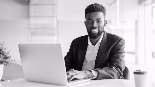 A smiling man in a blazer seated at a laptop in a bright office.